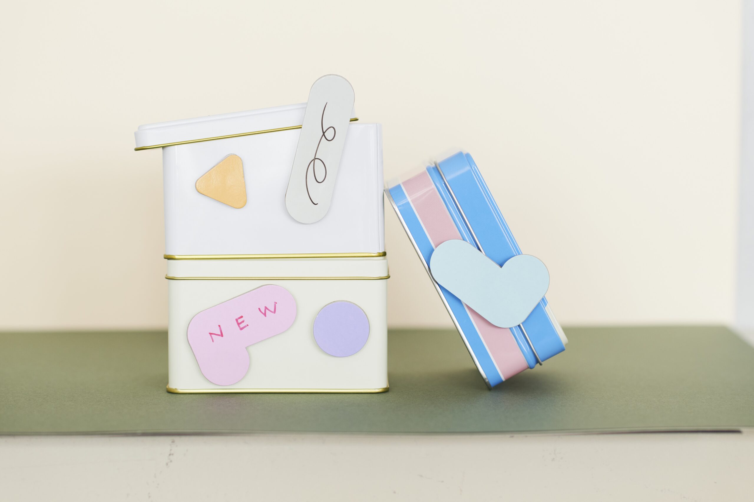 Stack of white tins with gold trim decorated by pastel stickers and a pale tag, a striped blue-pink craft item leaning against them.