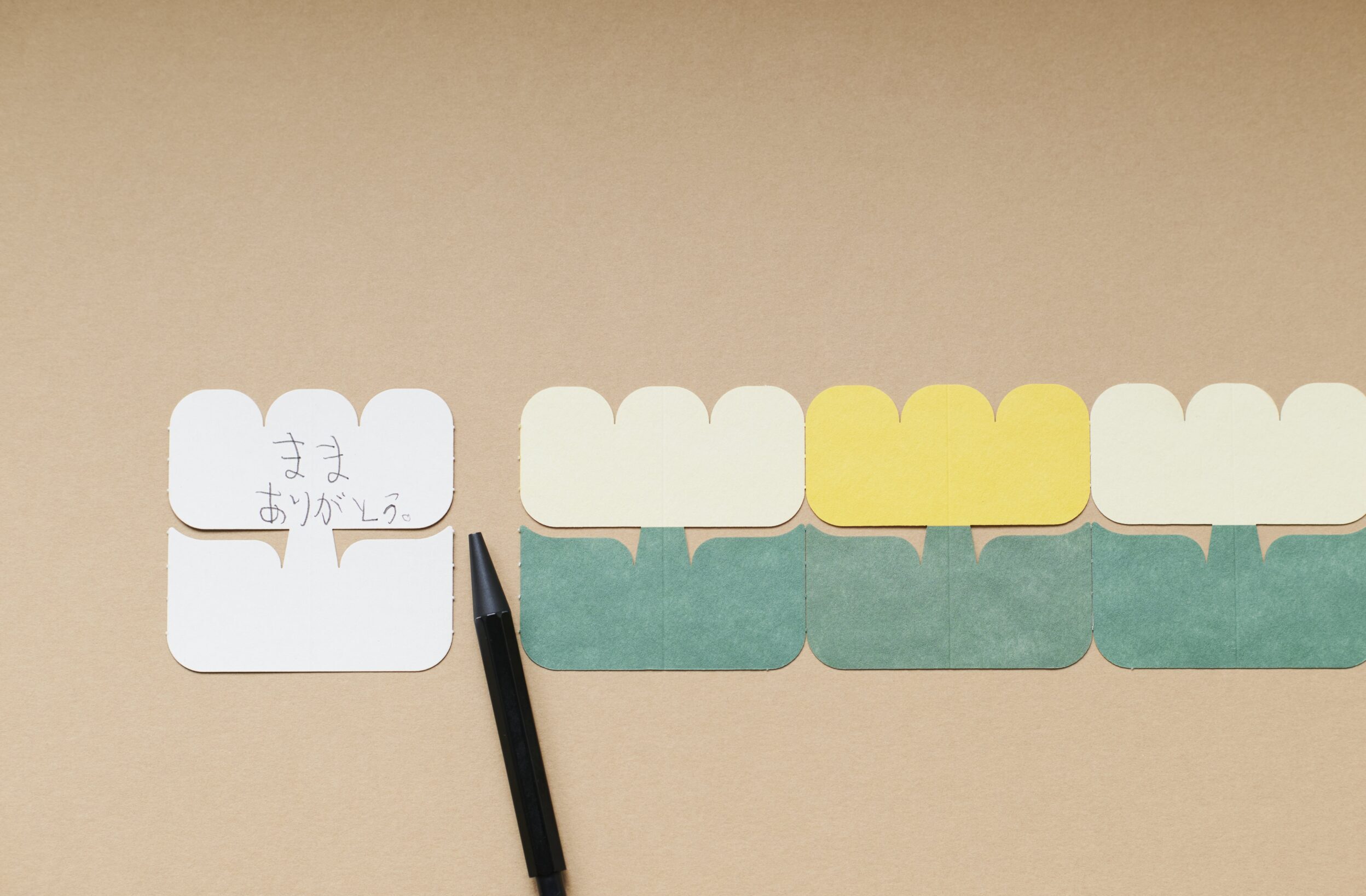 Row of rounded pastel color swatches along a brown paper background, with a black pencil and a small white card bearing handwritten Japanese text ありがとう.