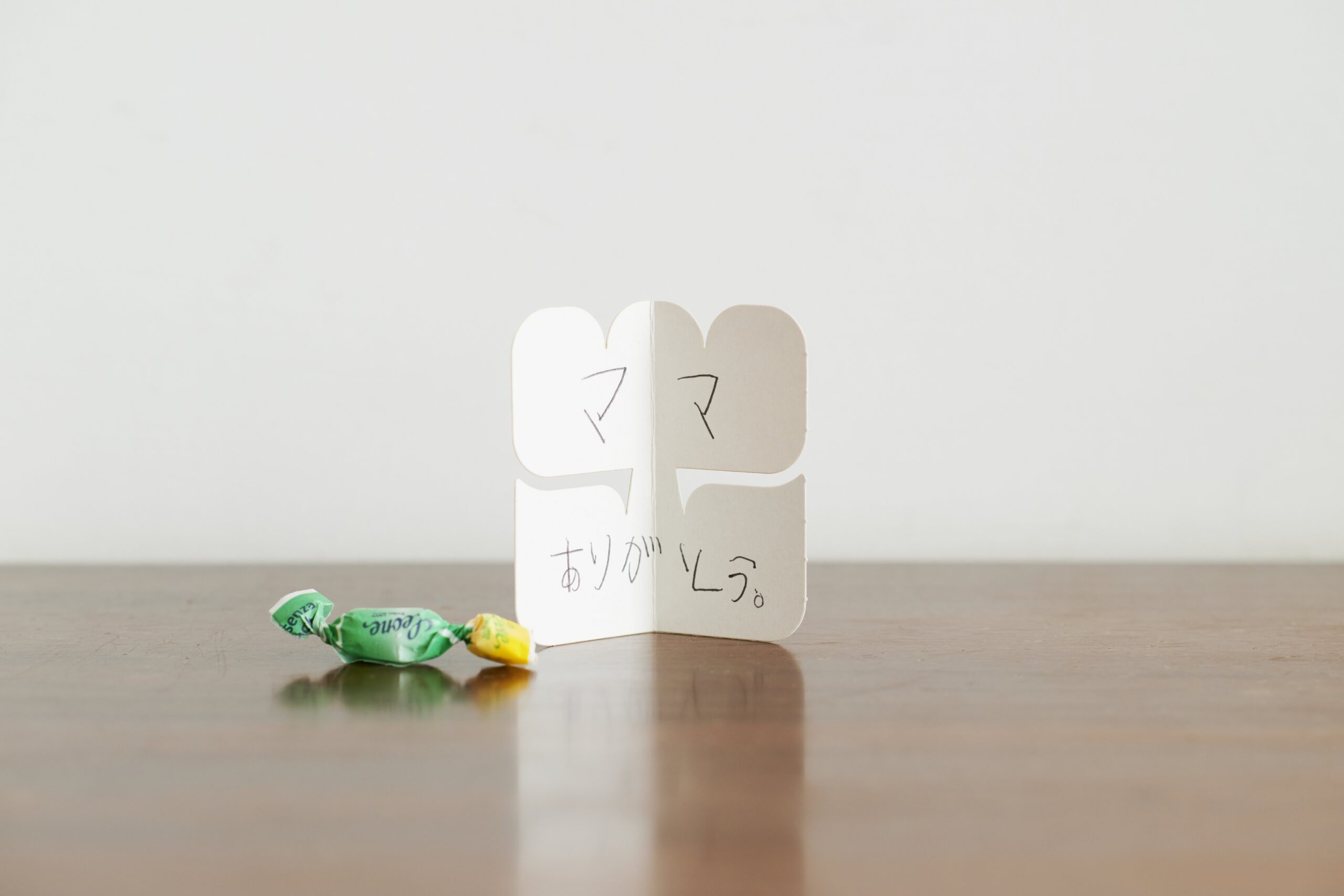 White double-heart shaped card standing on a wooden table, with the handwritten Japanese word ありがとう, and a crumpled green candy wrapper nearby.