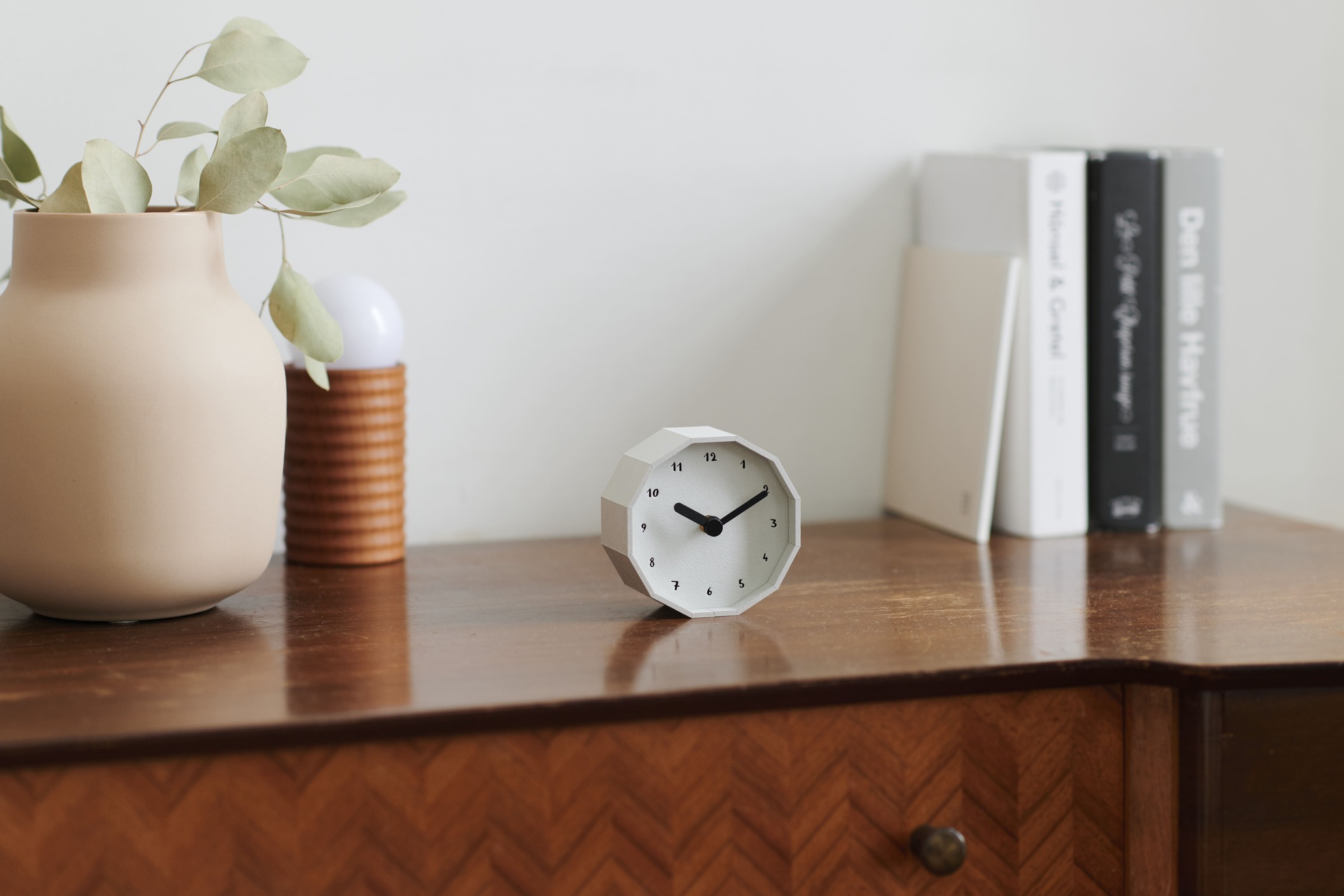 Desk setup with a beige ceramic vase containing eucalyptus, a geometric white clock, a ribbed brown vase, and a row of books on the right.