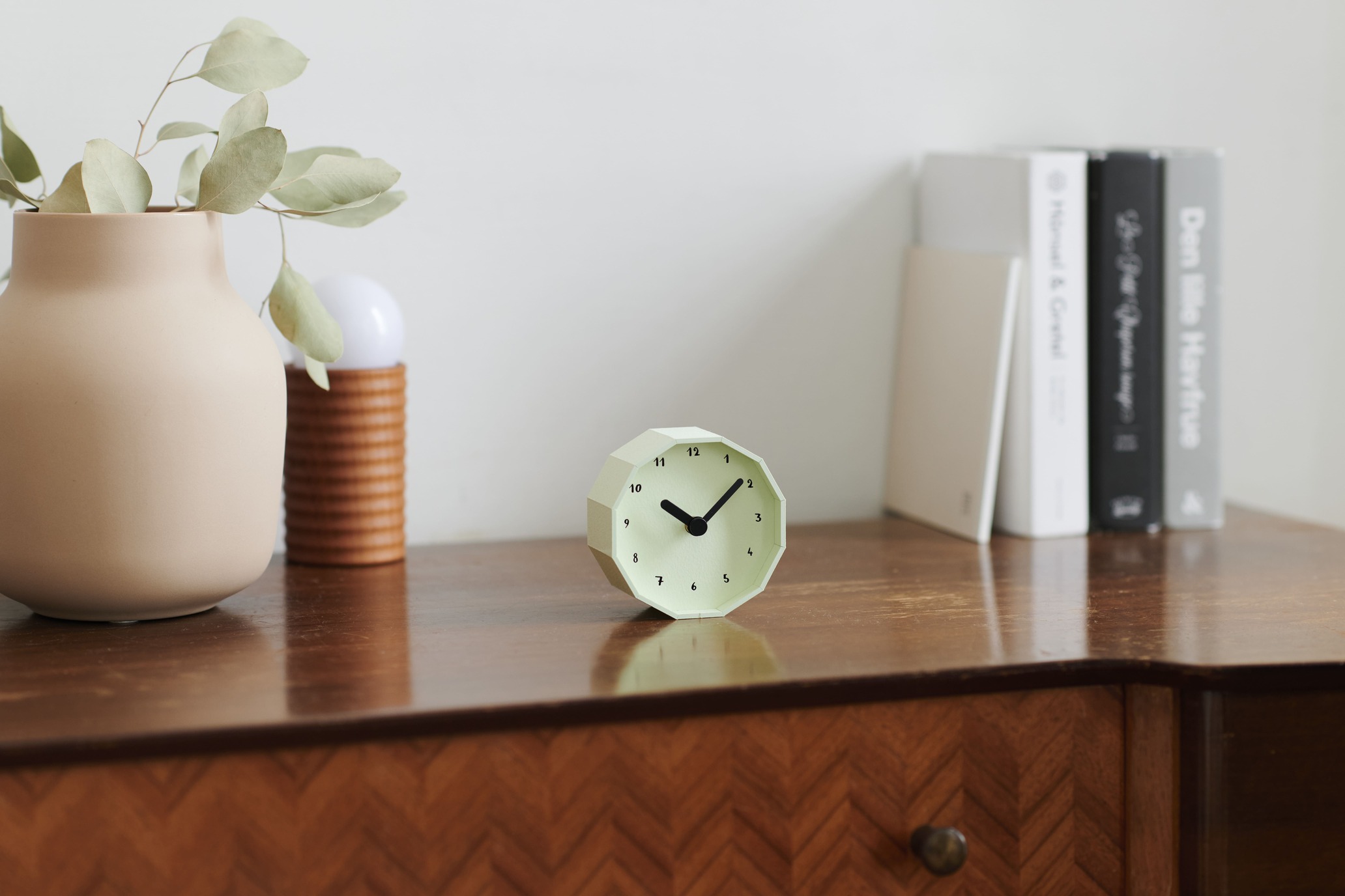 Decorative desk scene with a beige vase holding eucalyptus, a small mint-green geometric clock, and a row of books on wood.