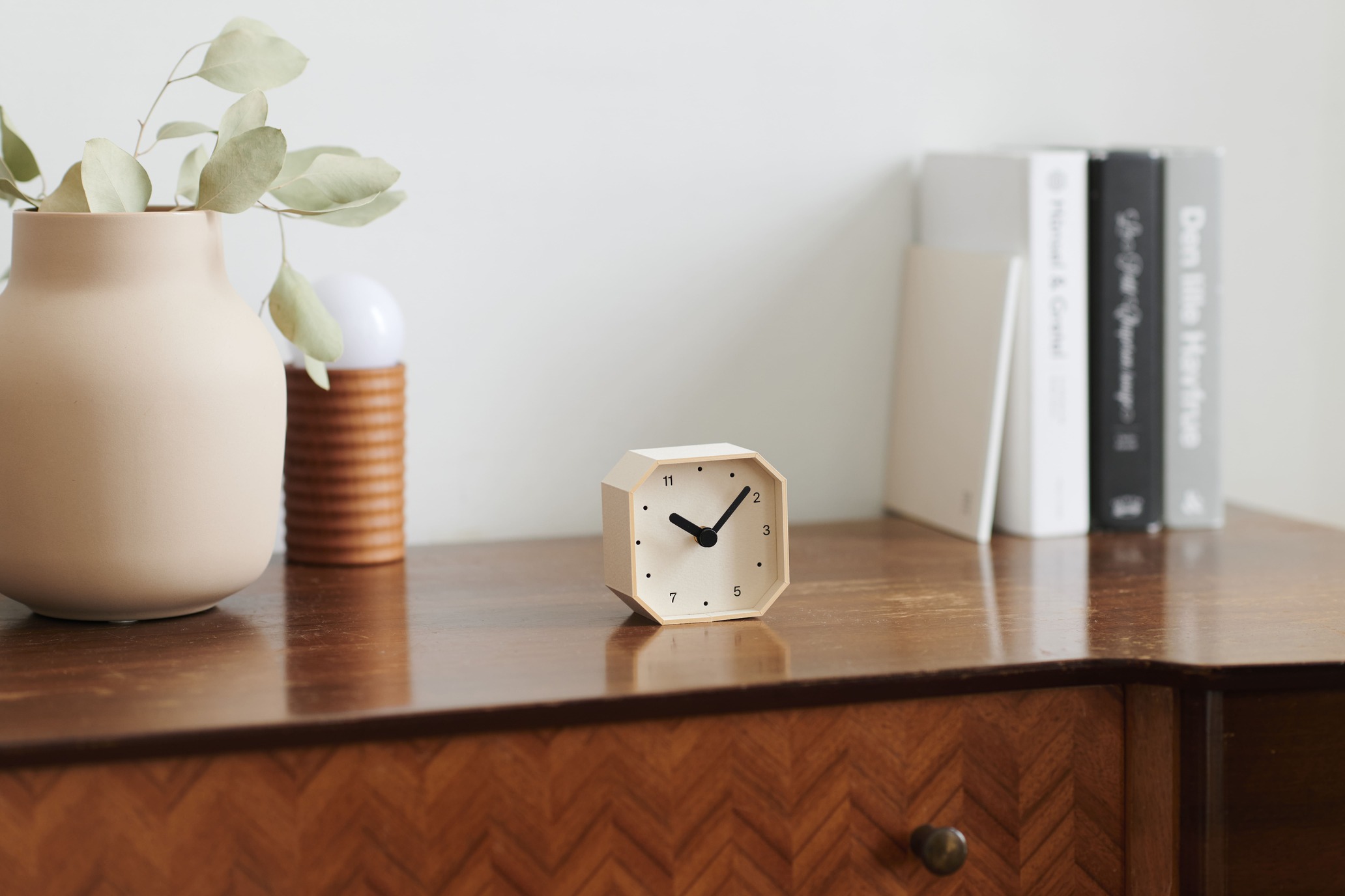 Beige ceramic vase with pale green eucalyptus on a wooden desk, a small geometric clock, and a row of books in the background.
