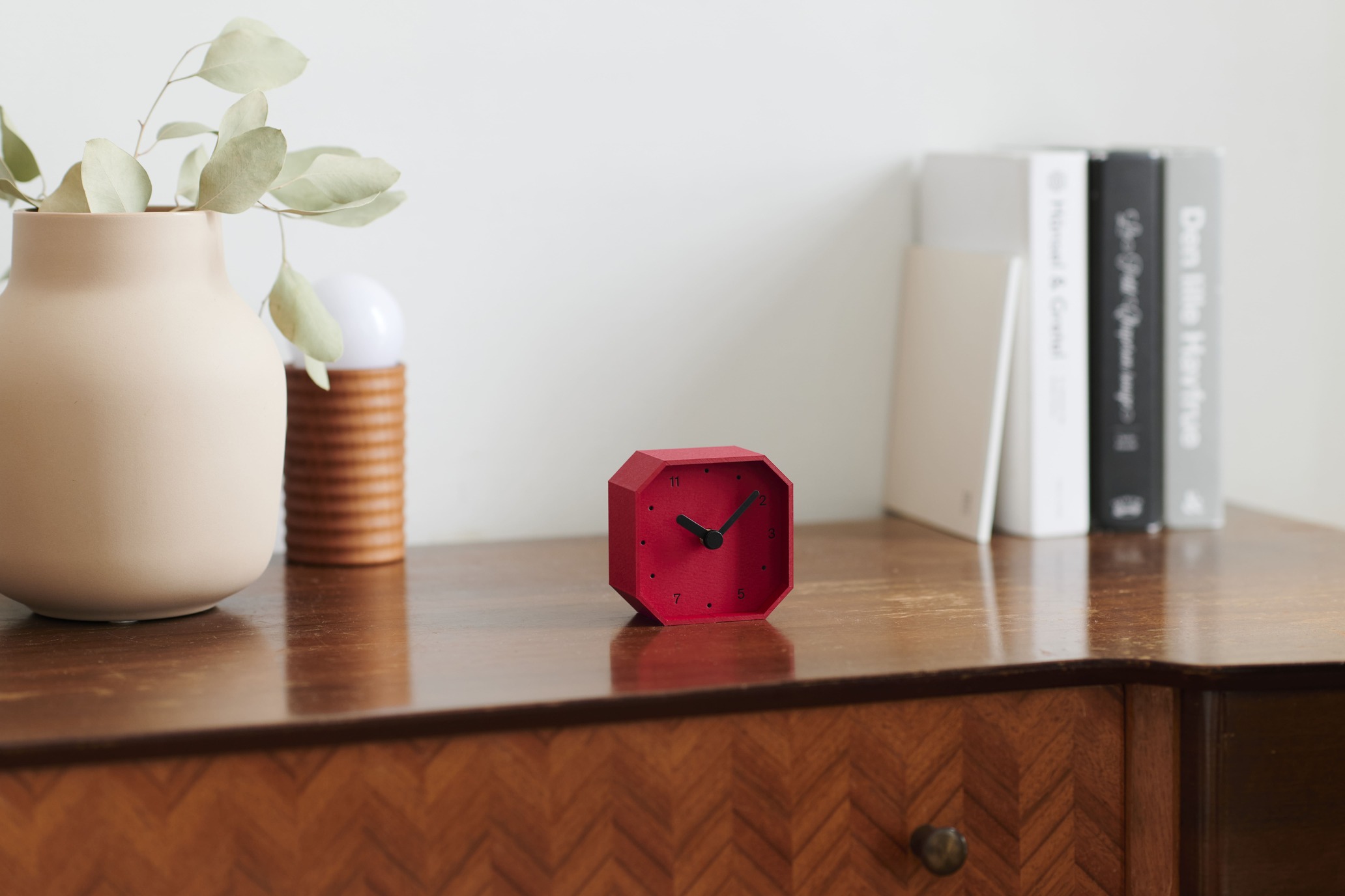 Red octagonal clock sits on a wooden desk beside a beige vase with green leaves and a row of books on the right.