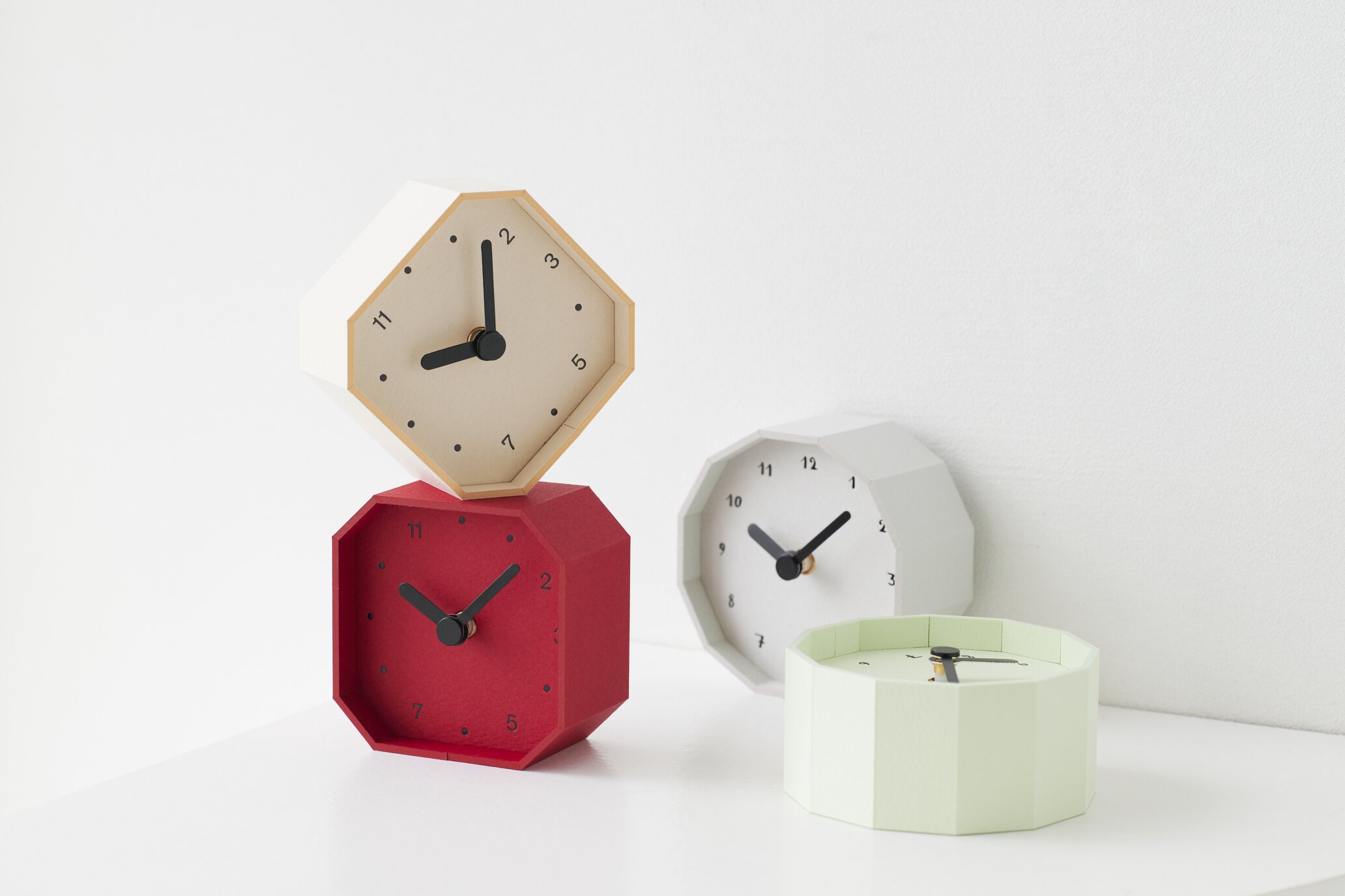 Stacked pastel hexagonal clocks in red, beige, and white sits on a white surface against a pale background, showing black clock hands.