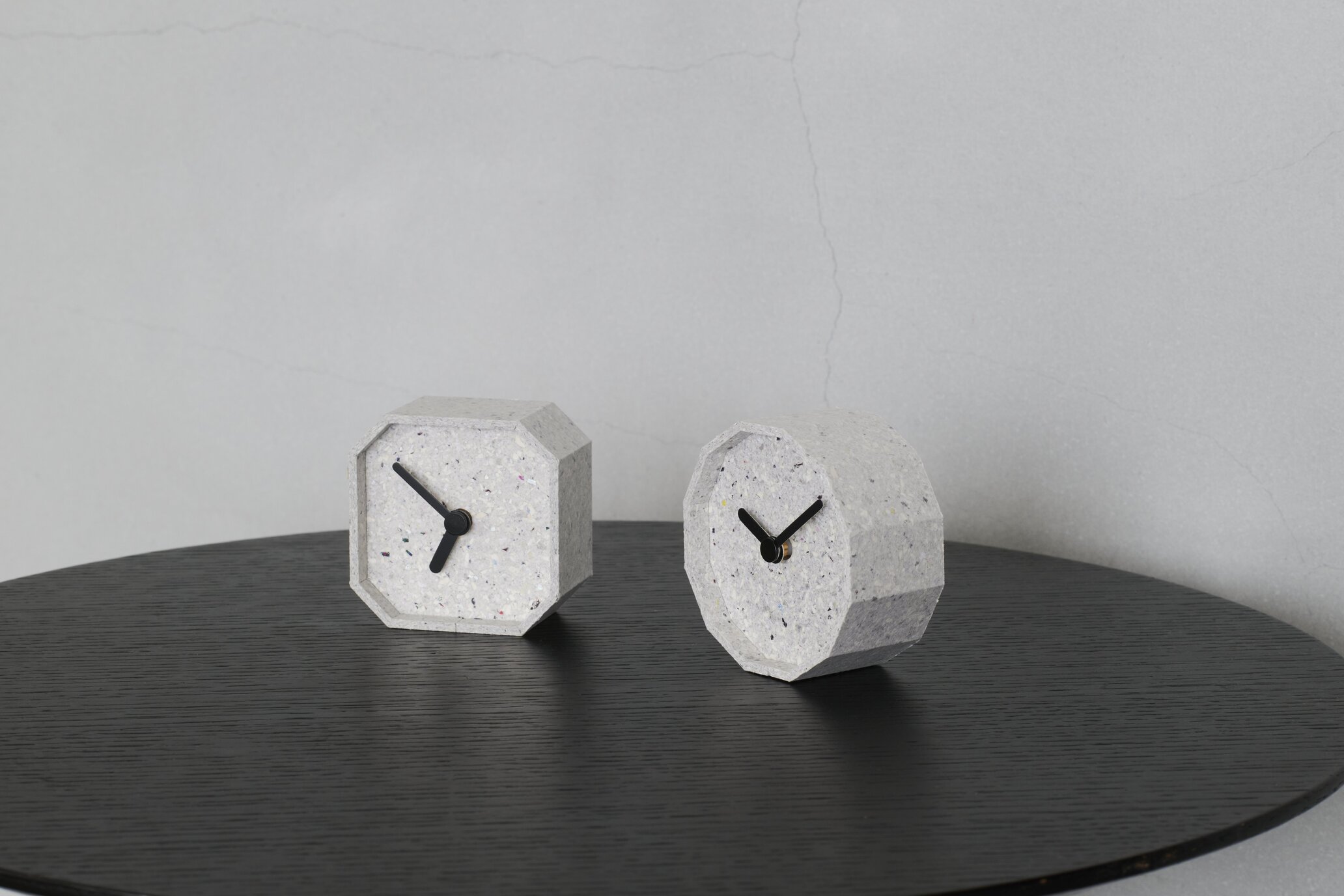 Two geometric concrete clocks with black hands sit on a dark circular table against a gray concrete wall, decorative timepieces.
