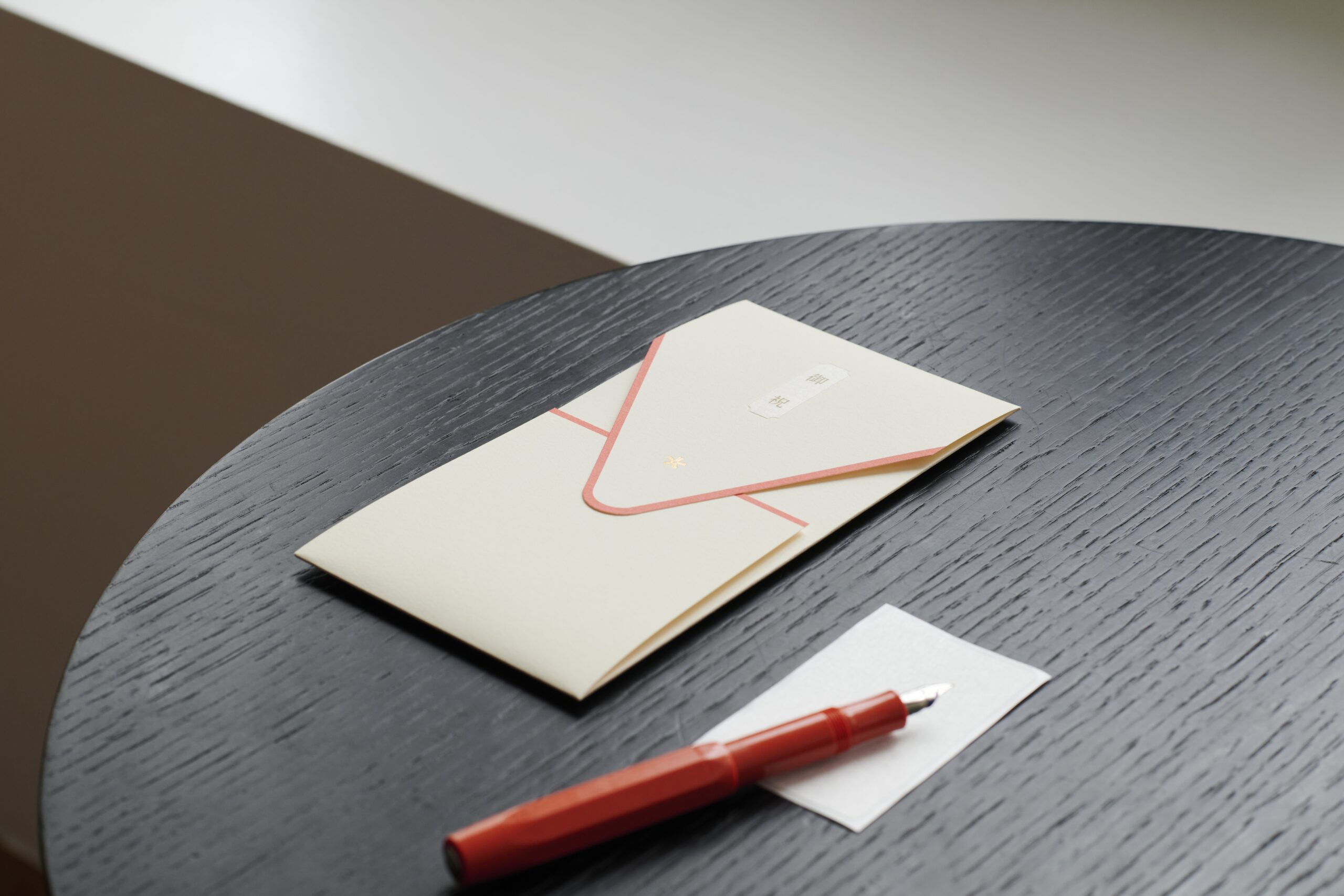 Red fountain pen resting on a pale envelope/card on a dark round table.