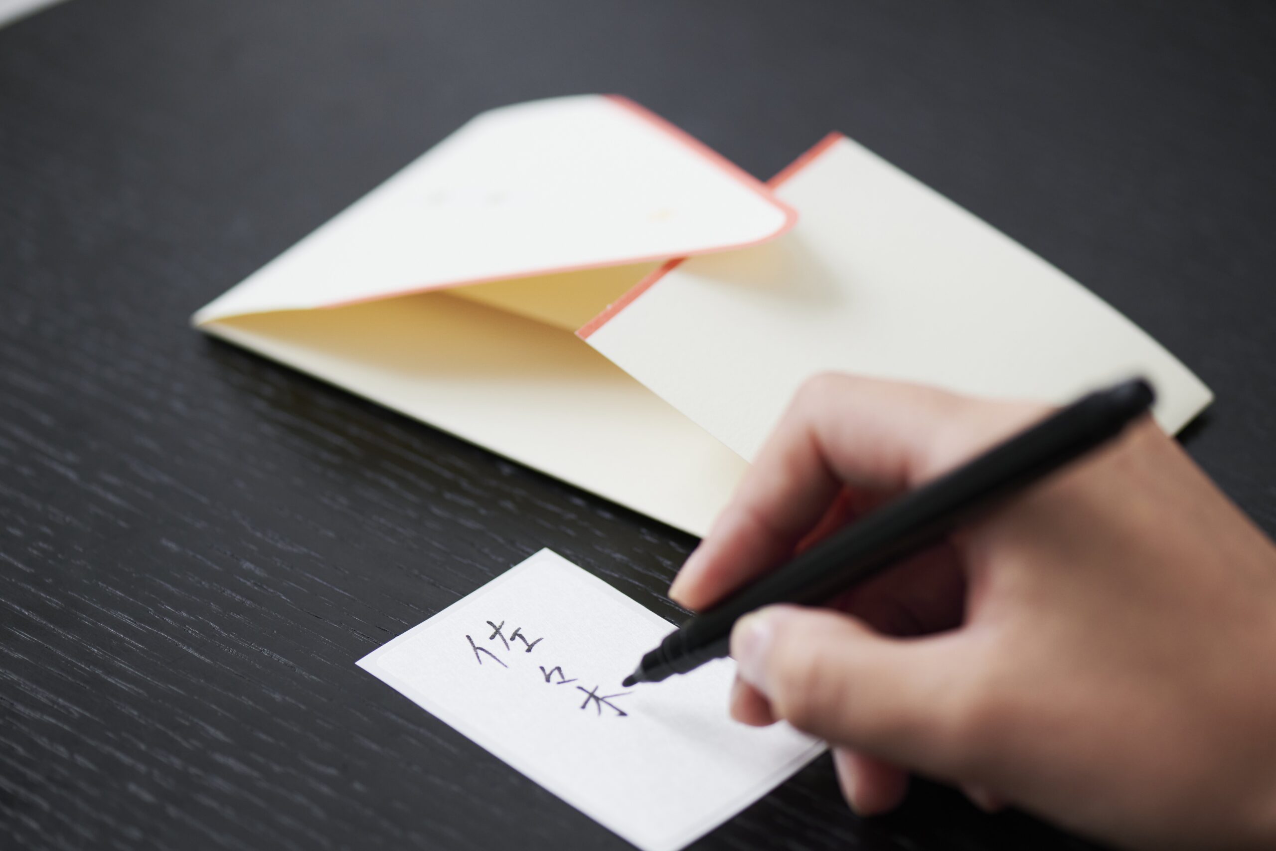 Close-up of a hand writing on a small white sticky note with a black pen on a dark wooden desk.