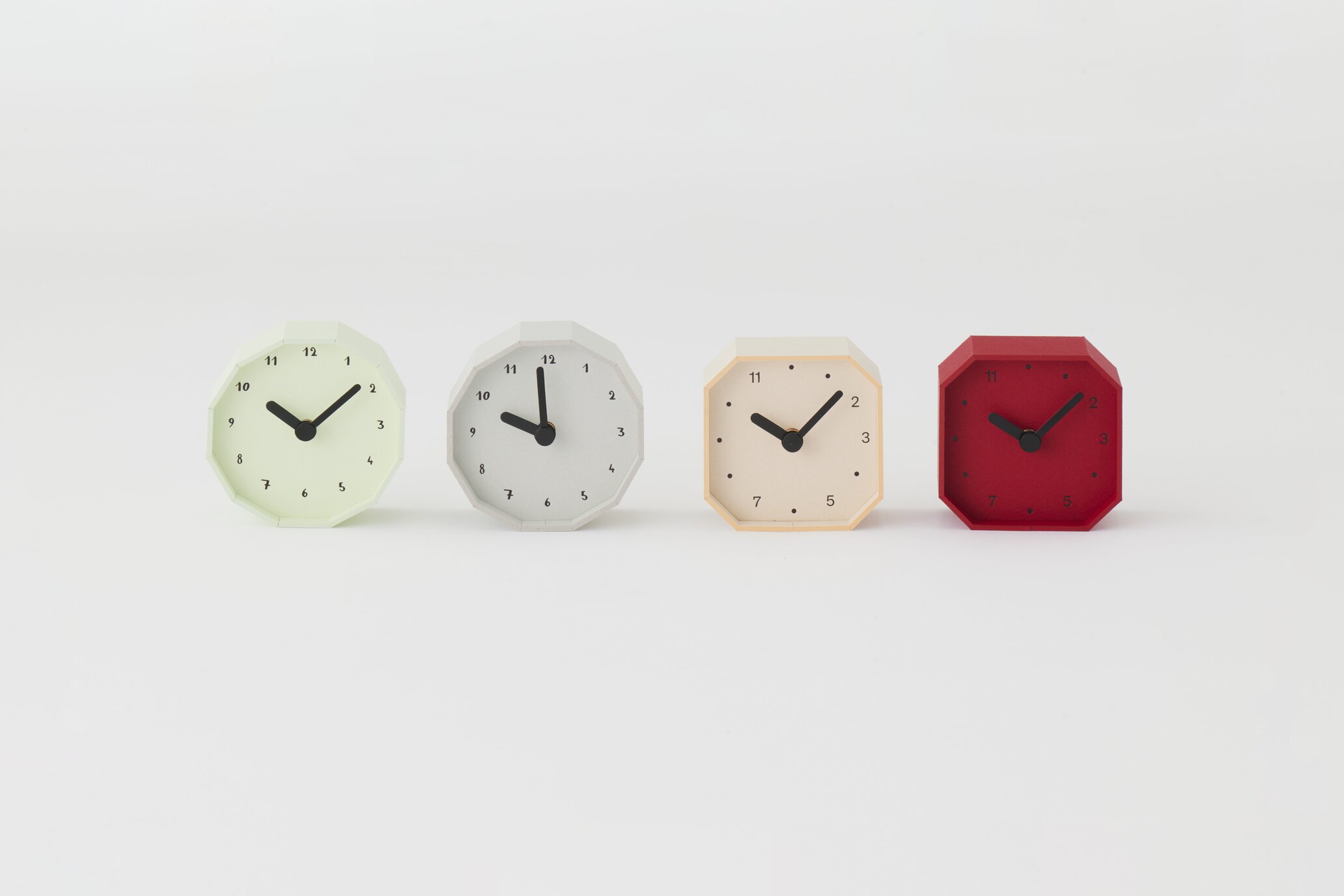 Four octagonal clocks in pastel colors (green, gray, beige, red) lined up on a white surface against a plain background.