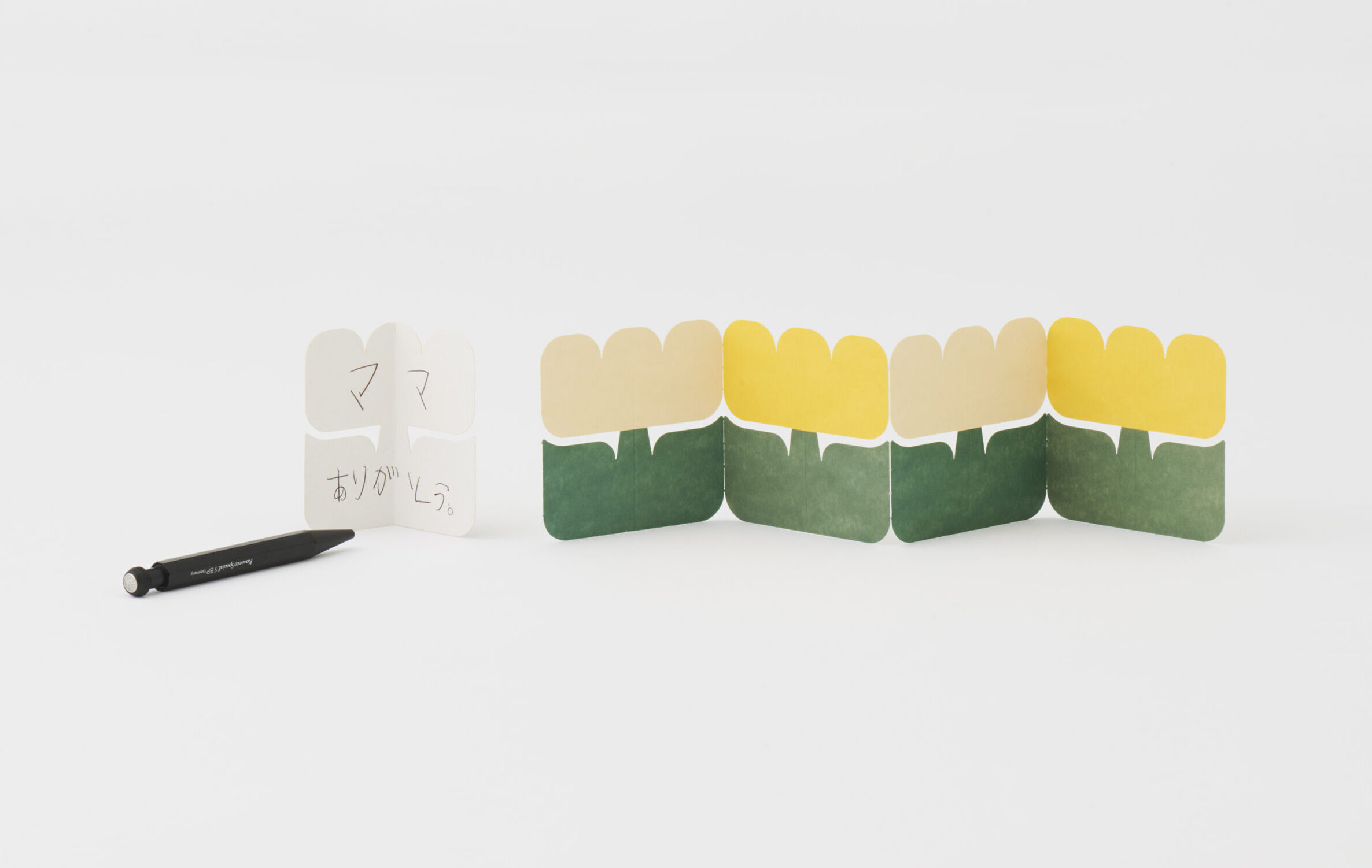 Row of green and yellow folded paper shapes lined up on a white surface, with a handwritten note and black marker to the left.